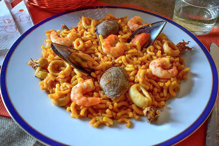 Plato de fideuá de marisco bien presentado, con los fideos melosos, los mariscos distribuidos, una cucharada de alioli al lado y una ramita de perejil fresco como decoración.